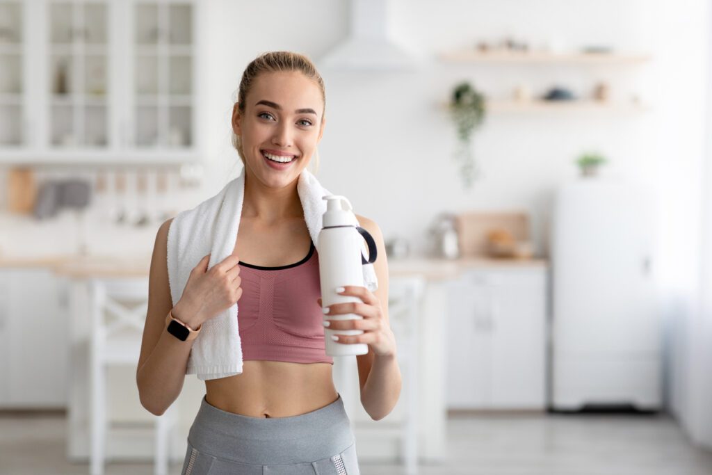 active sports at home and thirst, healthcare. Smiling young blonde woman in sportswear resting after workout in minimalist living room interior with towel on shoulders and bottle of water