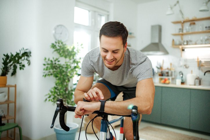 Young athletic man checking pulse on smart watch while riding bicycle in a domestic room at home